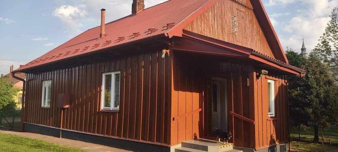 Czerwono-brązowy drewniany dom z czerwoną blachodachówką, widok z zewnątrz, mały pies na schodach wejściowych, w tle wieża kościelna.