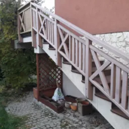 Zewnętrzne schody z drewnianą balustradą w kolorze beżowym, prowadzące do tarasu, z widoczną kamienną elewacją i dekoracyjnymi donicami u podstawy.
