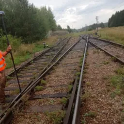 Pomiar geodezyjny na torach kolejowych; widoczne rozjazdy, osoba w kamizelce odblaskowej z tyczką geodezyjną, w tle zieleń i pochmurne niebo.