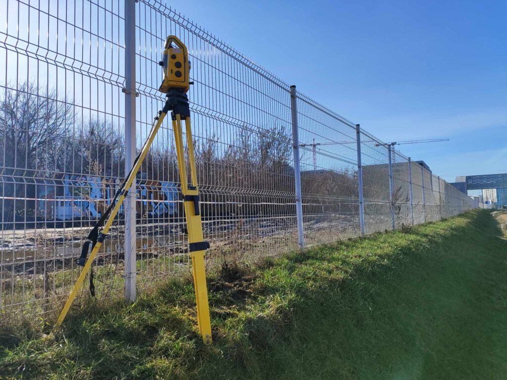 Żółty teodolit geodezyjny ustawiony na trójnogu, obok ogrodzenia z siatki, w tle budynek i drzewa.