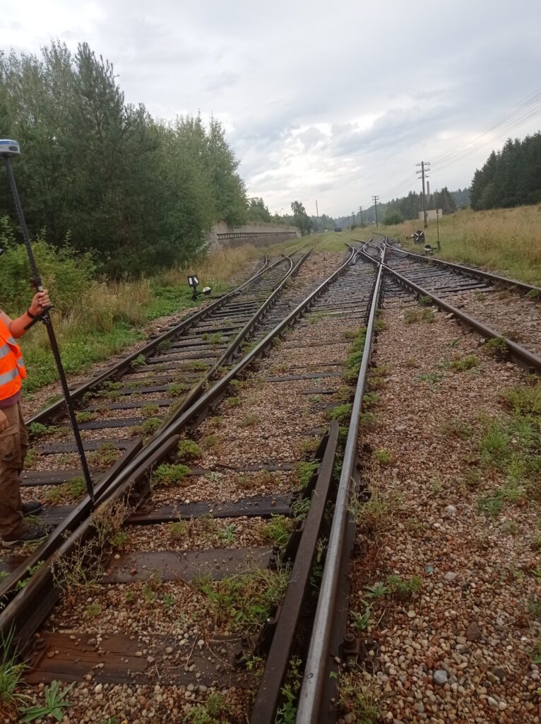 Pomiar geodezyjny na torach kolejowych; widoczne rozjazdy, osoba w kamizelce odblaskowej z tyczką geodezyjną, w tle zieleń i pochmurne niebo.