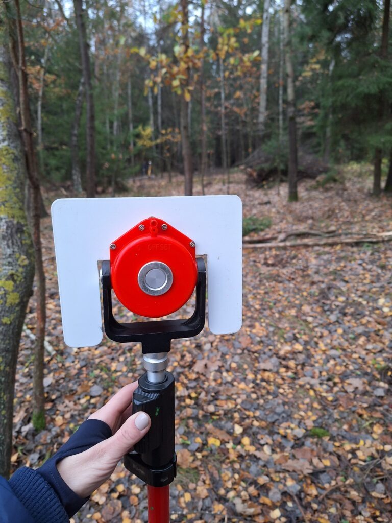 Pomiar geodezyjny w terenie leśnym z użyciem tyczki z reflektorem pryzmatycznym; widoczne opadłe liście i drzewa w tle.