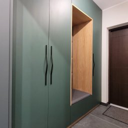 STOLMAX Meble SP ZOO SP K - Zielona szafa wnękowa z drewnianą wnęką do siedzenia, czarne podłużne uchwyty, szara poduszka siedziska i szara podłoga z płytek.