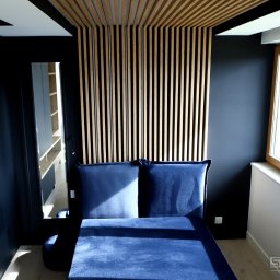 STOLMAX Meble SP ZOO SP K - Nowoczesna sypialnia z ciemnymi ścianami, drewnianym panelem dekoracyjnym na ścianie i suficie oraz niebieskim tapicerowanym łóżkiem z zagłówkiem.