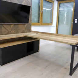 Nowoczesne biurko narożne z drewnianym blatem i czarnymi nogami, zintegrowane z czarną szafką z szufladami, na tle ściany z dekoracyjnym panelem i telewizorem.