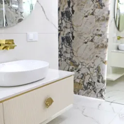 Nowoczesna łazienka z umywalką nablatową, złotą armaturą i marmurowym wykończeniem ścian oraz podłogi, widoczna szafka pod umywalkę z ryflowanym frontem i złotymi uchwytami.