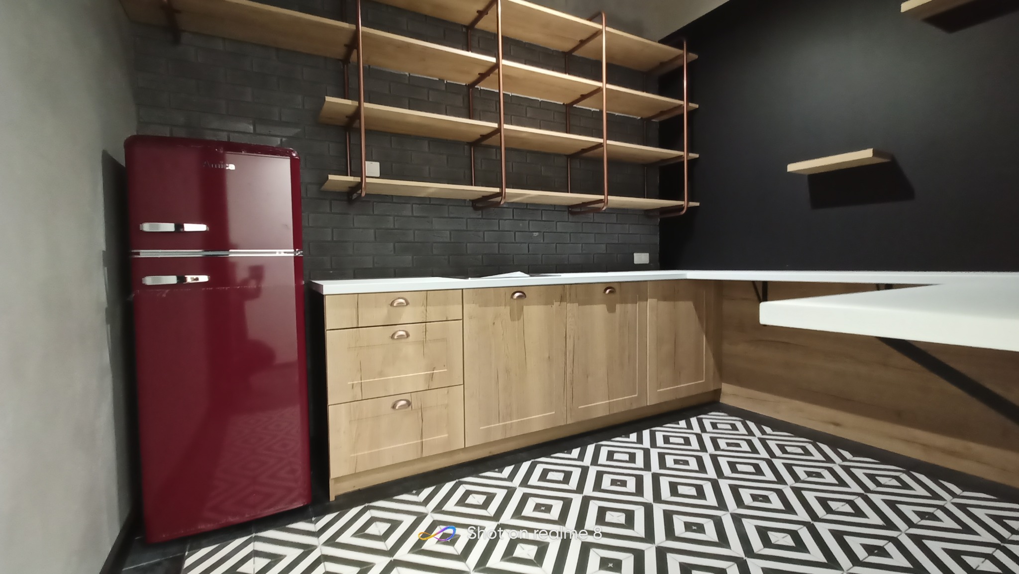 Nowoczesna kuchnia z drewnianymi szafkami, białym blatem, otwartymi półkami z miedzianymi wspornikami, czarną ceglaną ścianą i czerwona lodówką w stylu retro oraz podłogą w geometryczny wzór.