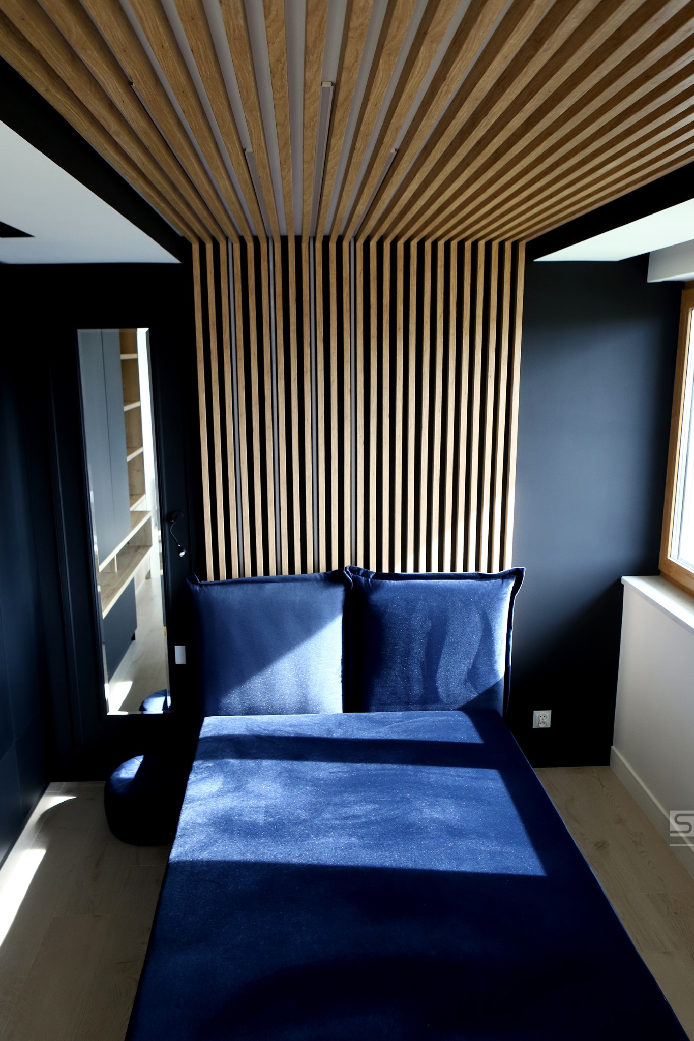 Nowoczesna sypialnia z ciemnymi ścianami, drewnianym panelem dekoracyjnym na ścianie i suficie oraz niebieskim tapicerowanym łóżkiem z zagłówkiem.