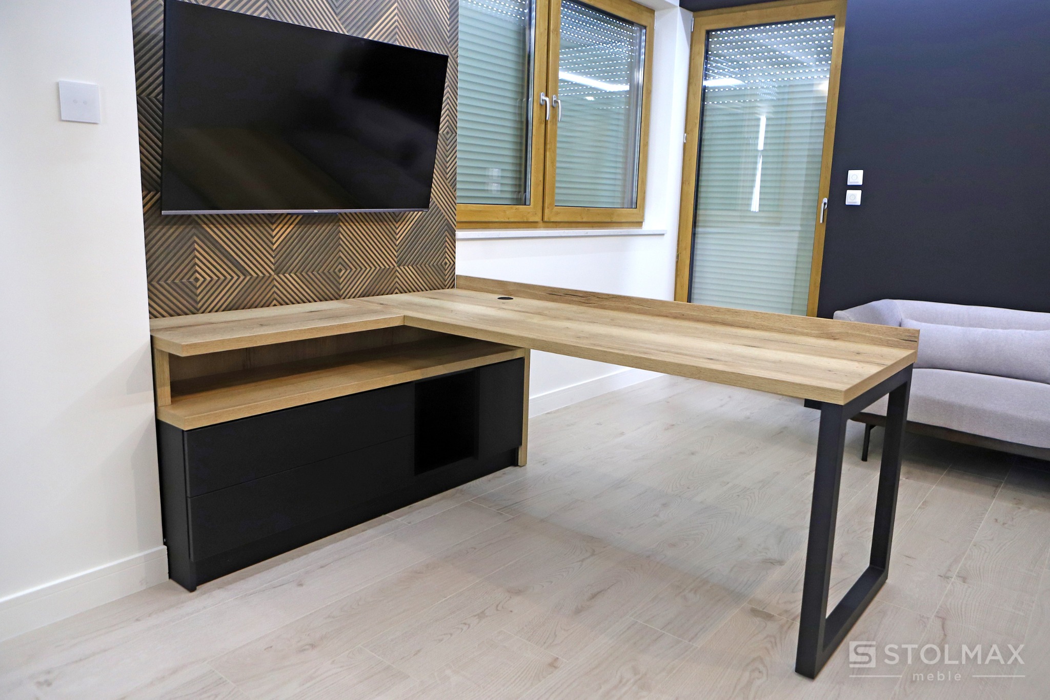 Nowoczesne biurko narożne z drewnianym blatem i czarnymi nogami, zintegrowane z czarną szafką z szufladami, na tle ściany z dekoracyjnym panelem i telewizorem.