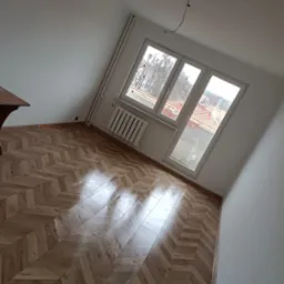 Jasny pokój po remoncie z nową podłogą ułożoną w jodełkę, widok na okno z grzejnikiem i fragment stołu, w tle zabudowa miejska.