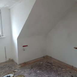 Akacjowa - Pokój na poddaszu w trakcie prac wykończeniowych: ściany pokryte świeżą warstwą gładzi, widoczne puszki instalacji elektrycznej, na podłodze resztki materiałów i puszka farby.