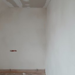 Akacjowa - Fragment wnętrza po pracach wykończeniowych: biała ściana z gładzią, widoczne ślady po instalacji elektrycznej oraz fragment listwy przypodłogowej.