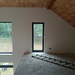 Akacjowa - Wnętrze poddasza z drewnianą boazerią na suficie, świeżo pomalowane na biało ściany, aluminiowa drabina leżąca na betonowej posadzce, duże okna z widokiem na zieleń.