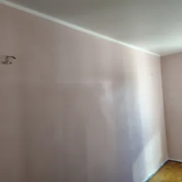 Świeżo pomalowana ściana w odcieniach pudrowego różu z widocznymi śladami po zamontowanym wcześniej uchwycie, listwa przypodłogowa i zabezpieczona taśmą podłoga parkietowa.