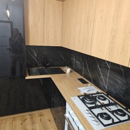 Akacjowa - Nowoczesna kuchnia z drewnianymi szafkami, czarnym blatem i marmurową ścianą. Widoczna płyta gazowa i piekarnik. W odbiciu szyby widać osobę robiącą zdjęcie.