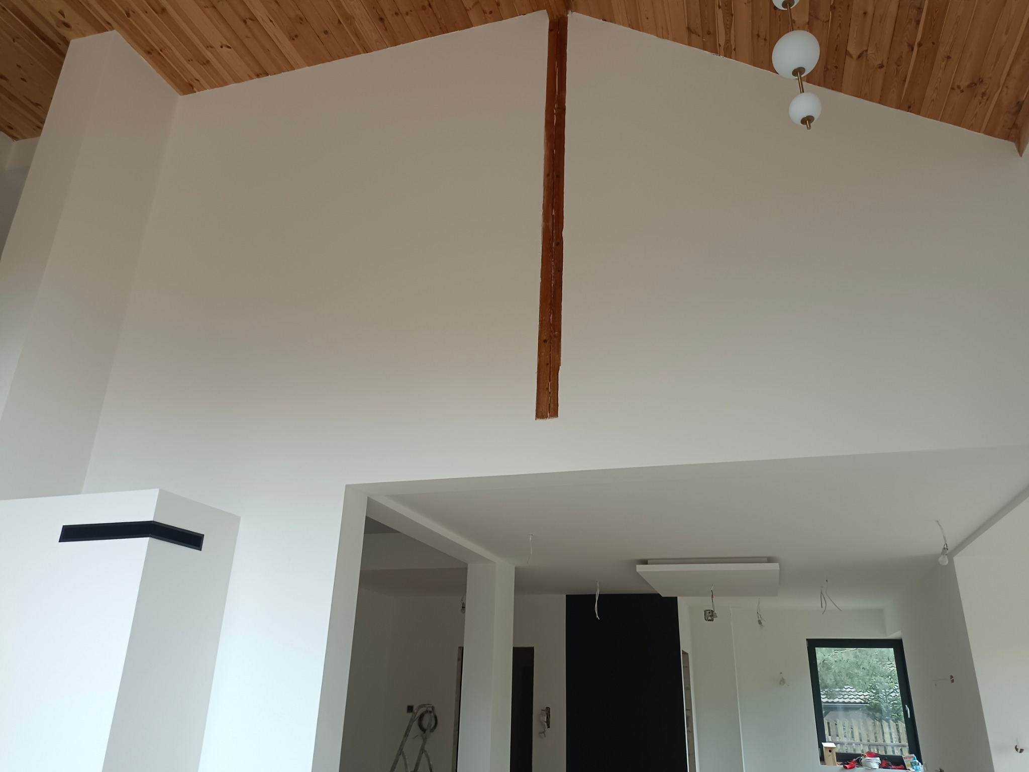 Wnętrze po malowaniu z wysokim sufitem i drewnianą boazerią, widoczny fragment drewnianej belki konstrukcyjnej oraz przygotowane punkty oświetleniowe.
