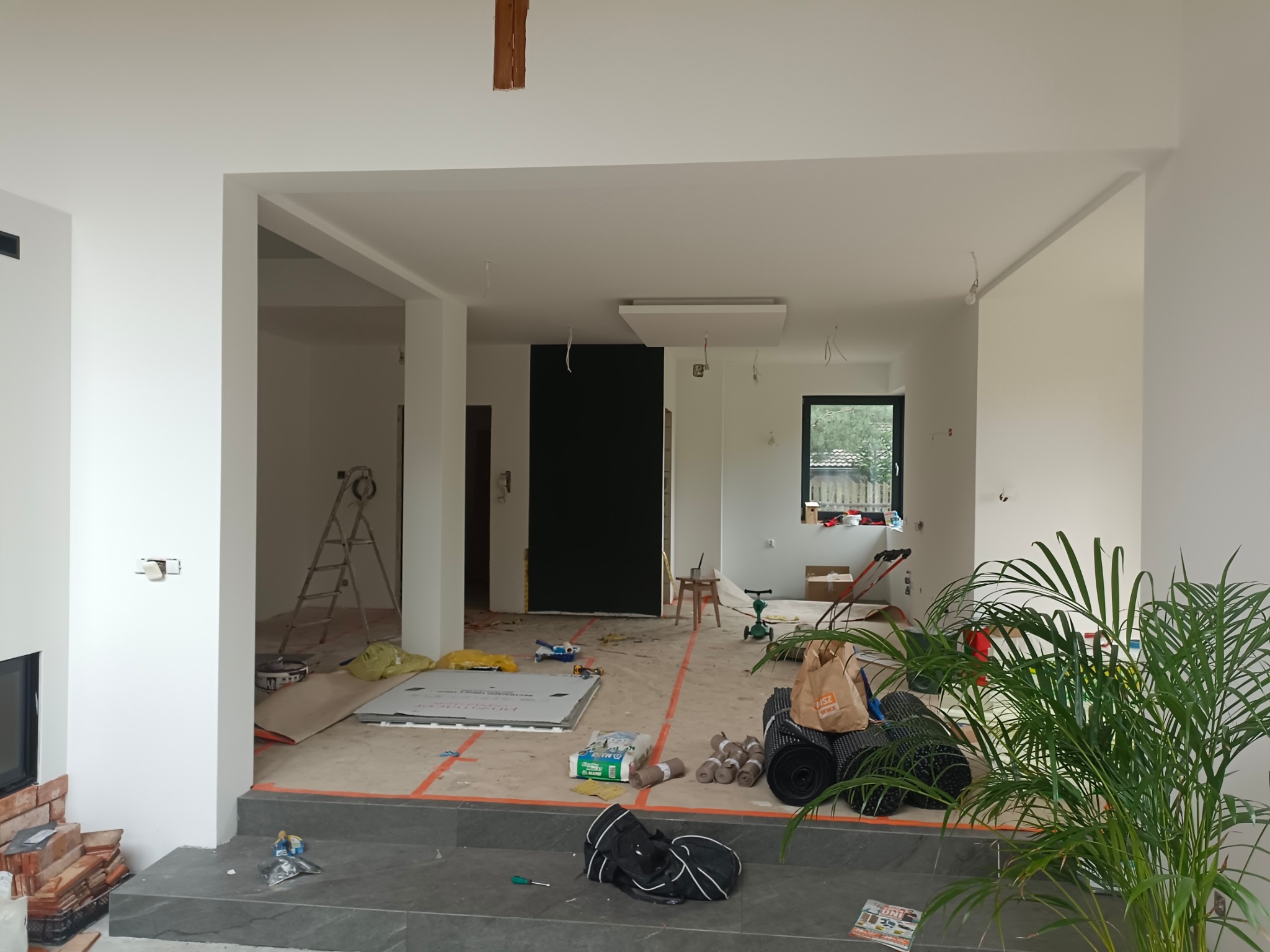 Wnętrze w trakcie remontu: świeżo pomalowane ściany na biało, widoczne przygotowania do układania podłogi, rozłożone materiały budowlane, drabina i narzędzia, roślina doniczkowa na pierwszym planie.