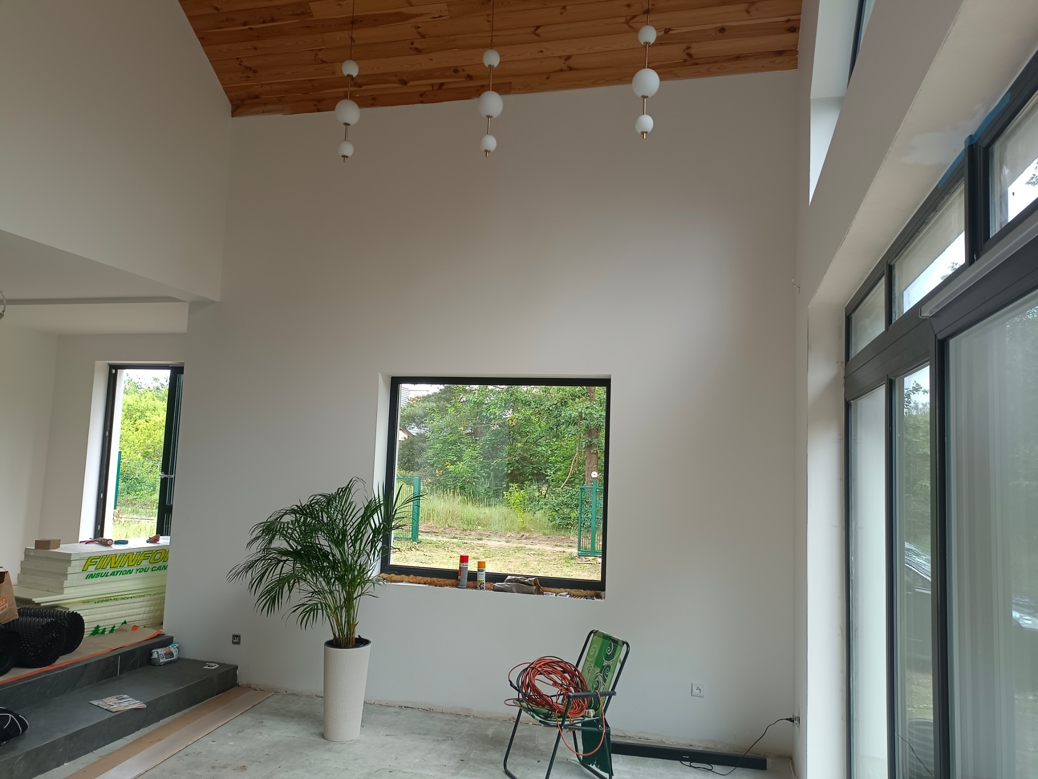 Wnętrze w trakcie wykończenia: białe ściany, drewniany sufit z nowoczesnym oświetleniem, okna z czarnymi ramami, roślina doniczkowa, materiały izolacyjne i krzesło z kablem elektrycznym...