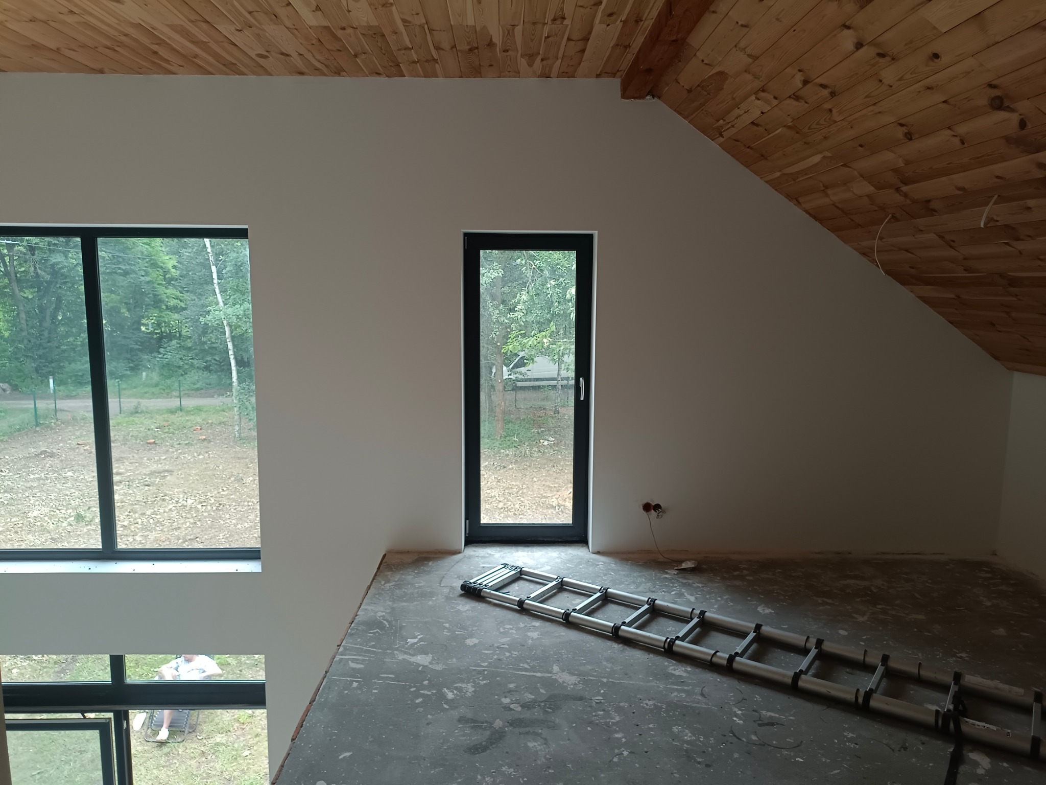 Wnętrze poddasza z drewnianą boazerią na suficie, świeżo pomalowane na biało ściany, aluminiowa drabina leżąca na betonowej posadzce, duże okna z widokiem na zieleń.