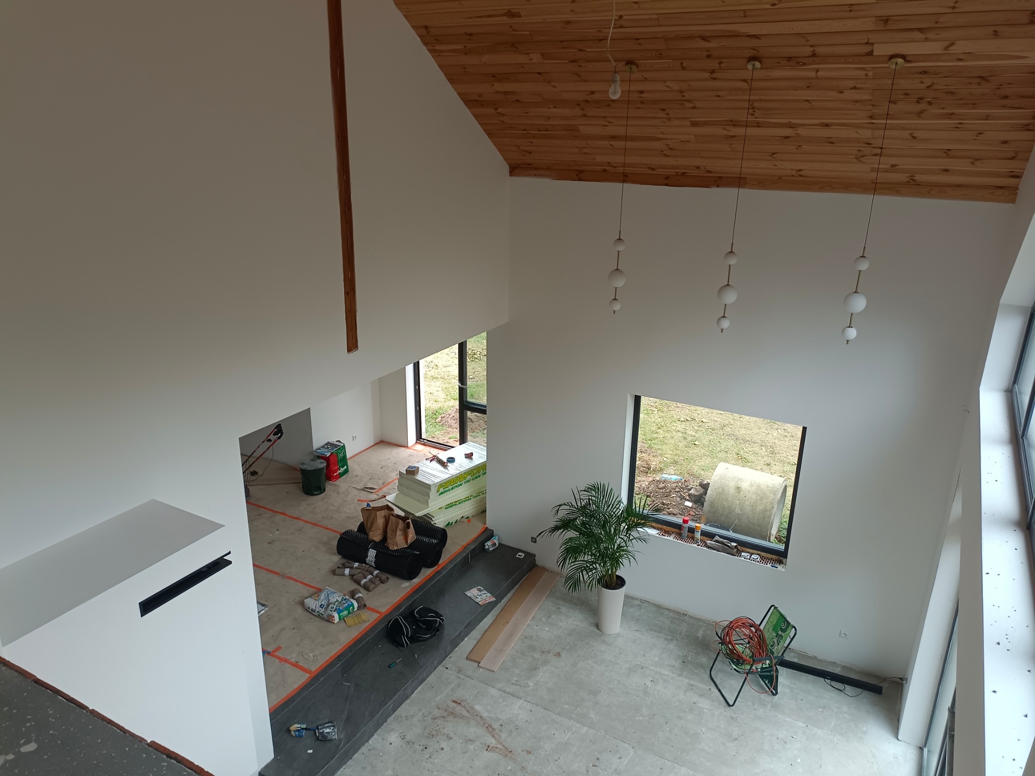 Wnętrze domu w trakcie prac wykończeniowych, widok z góry: białe ściany, drewniany sufit, podłoga przygotowana pod wylewkę, widoczne materiały budowlane, narzędzia i roślina doniczkowa.