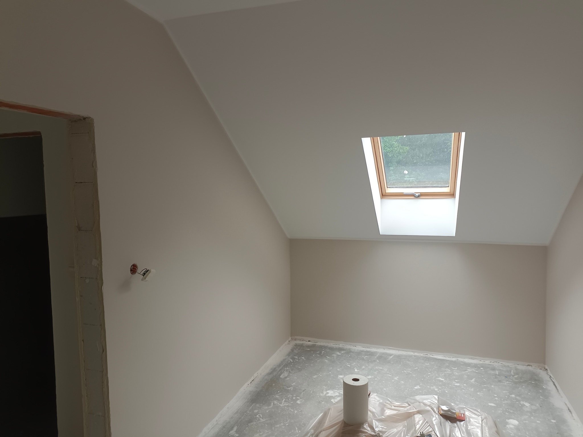 Pomalowane na beżowo poddasze ze skośnym sufitem i oknem dachowym; widoczne zabezpieczone folią podłoże, rolka do malowania i niedokończone obramowanie drzwi.