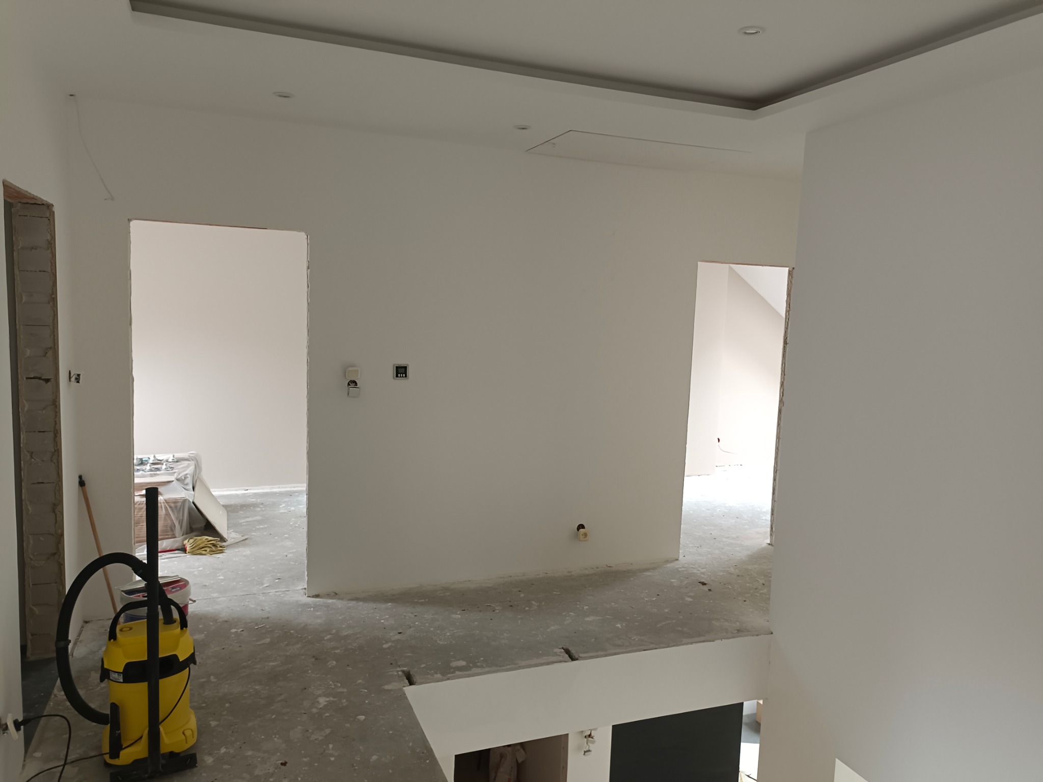 Wnętrze w trakcie prac wykończeniowych: świeżo pomalowane białe ściany, widoczny żółty odkurzacz przemysłowy, przygotowane podłoże pod panele, niezabezpieczone otwory drzwiowe i sufit z wnęką.