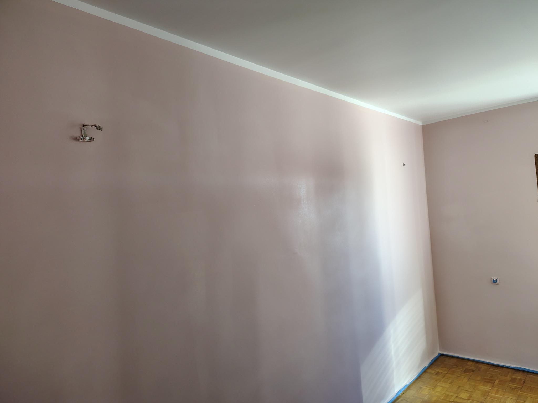Świeżo pomalowana ściana w odcieniach pudrowego różu z widocznymi śladami po zamontowanym wcześniej uchwycie, listwa przypodłogowa i zabezpieczona taśmą podłoga parkietowa.