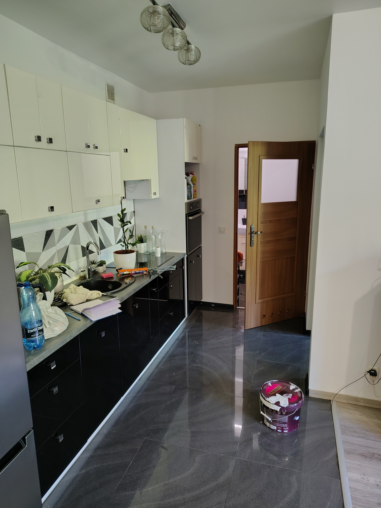 Kuchnia w trakcie wykończenia: czarne szafki, błyszcząca podłoga, pojemnik z farbą na podłodze, przygotowania do prac wykończeniowych.