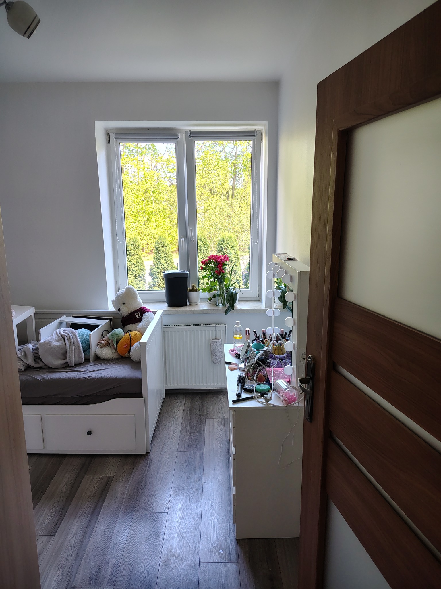 Jasny pokój z łóżkiem, biurkiem kosmetycznym i oknem z widokiem na zieleń. Drewniana podłoga i białe meble. Na łóżku pluszowy miś i poduszki.