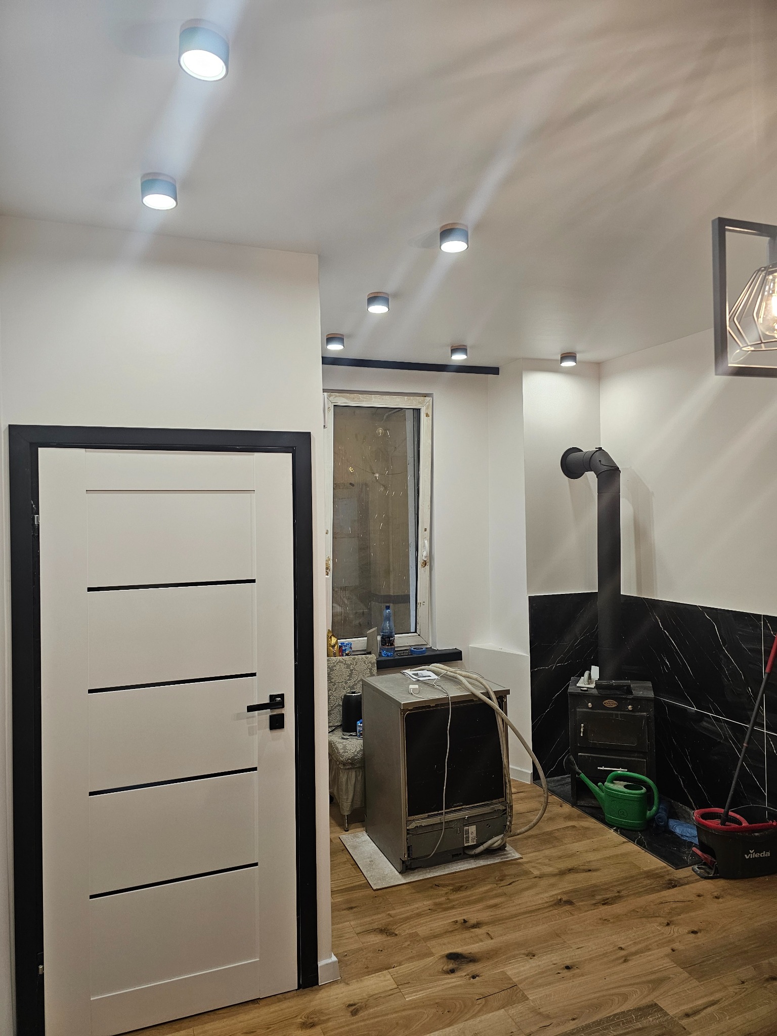 Jasne wnętrze po remoncie: białe ściany z czarnymi akcentami, drewniana podłoga, piec kaflowy i nowoczesne oświetlenie sufitowe. Widoczny sprzęt AGD w trakcie montażu.