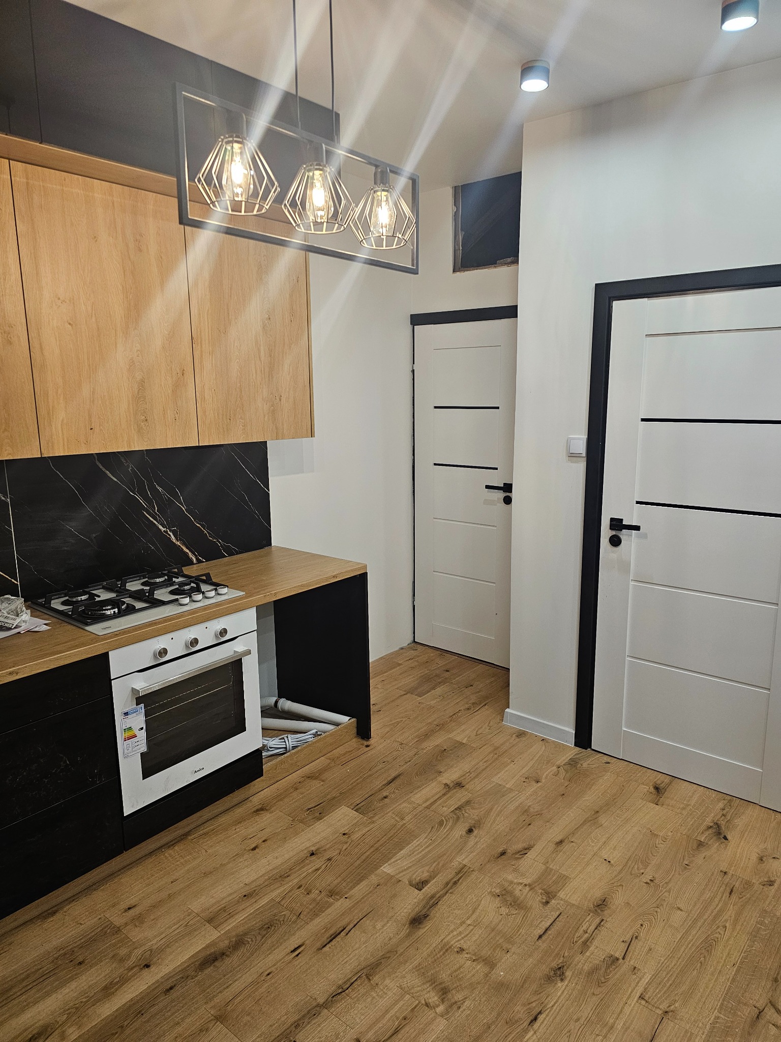 Nowoczesna kuchnia z drewnianymi szafkami, blatem imitującym drewno i czarnym marmurem na ścianie. Widoczne białe drzwi z czarnymi akcentami i drewniana podłoga.