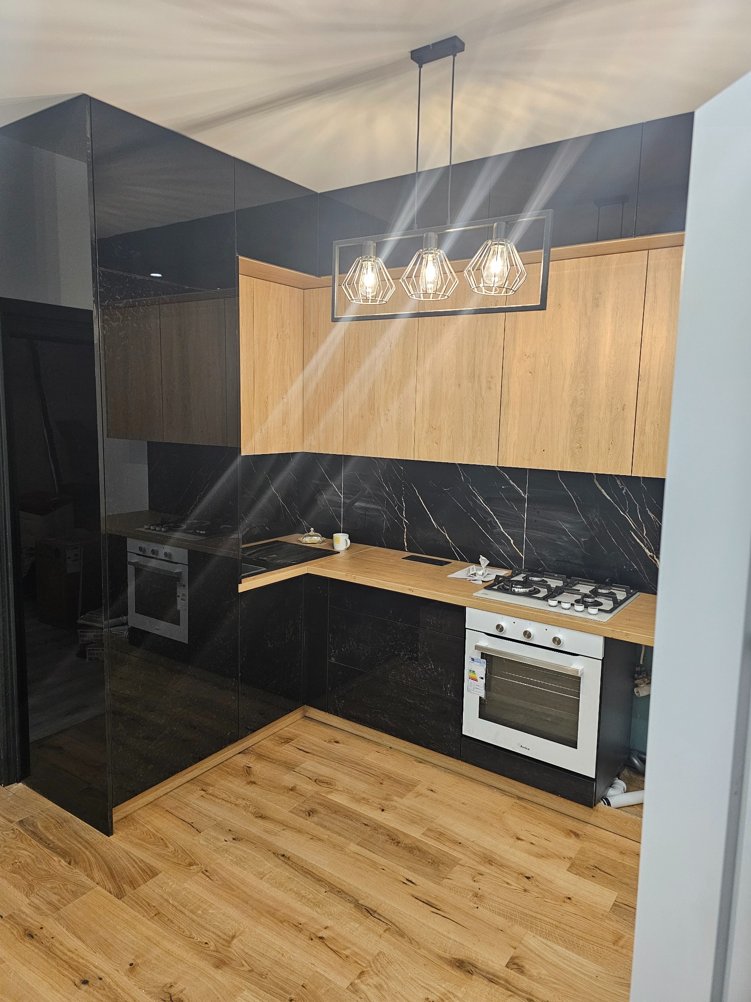 Nowoczesna kuchnia z drewnianymi szafkami, czarnym marmurem na ścianie i oryginalnym oświetleniem. Widoczne panele podłogowe i sprzęt AGD.