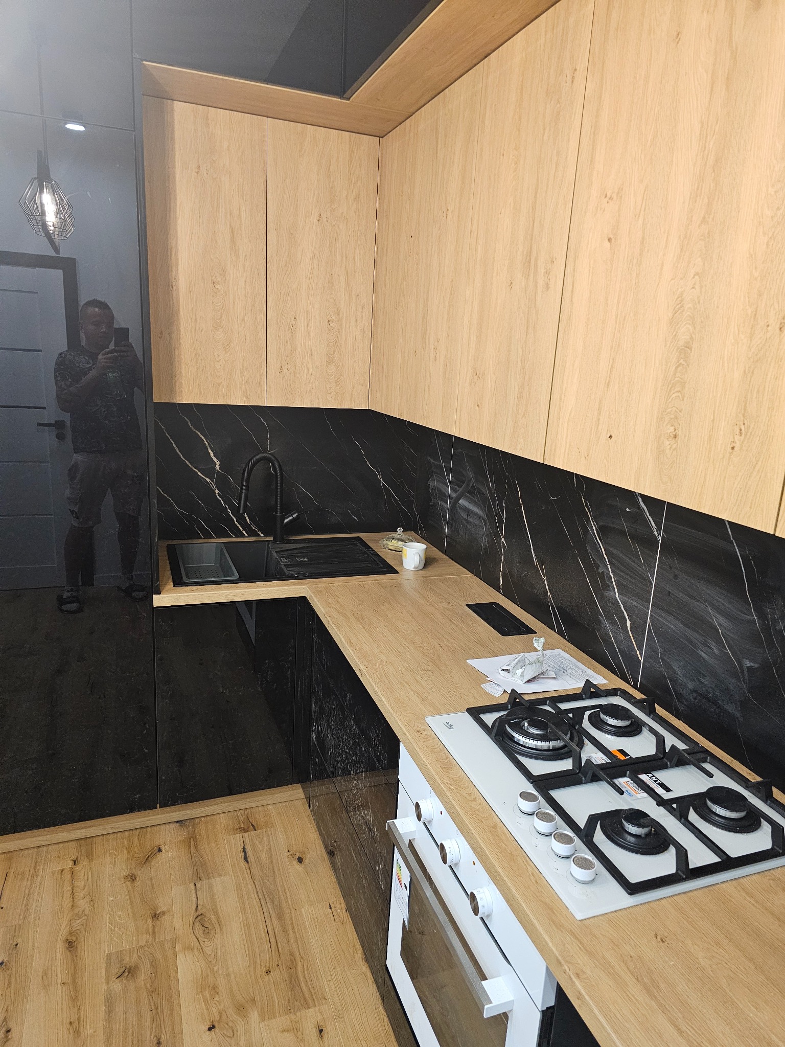 Nowoczesna kuchnia z drewnianymi szafkami, czarnym blatem i marmurową ścianą. Widoczna płyta gazowa i piekarnik. W odbiciu szyby widać osobę robiącą zdjęcie.