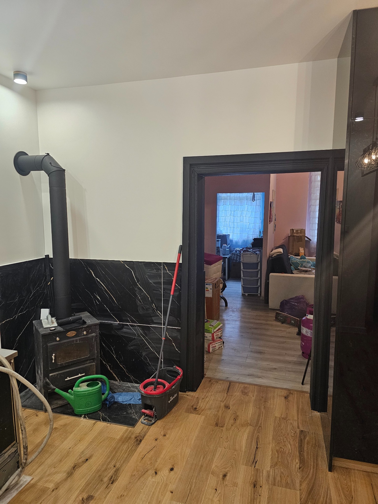 Wnętrze po remoncie: drewniana podłoga, czarny piec z marmurowym wykończeniem ściany. Widok na pokój przez czarne obramowanie drzwi. Mop i akcesoria do sprzątania.