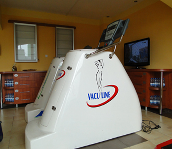 Dwa urządzenia Vacu Line w gabinecie, obok szafka z wodą, widoczny ekran. Na urządzeniach logo z sylwetką kobiety.