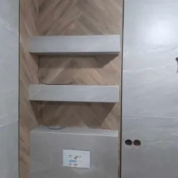 Wykończona ściana łazienki z drewnopodobną glazurą ułożoną w jodełkę, szare półki i zabudowa stelaża WC z instrukcją JOMO.