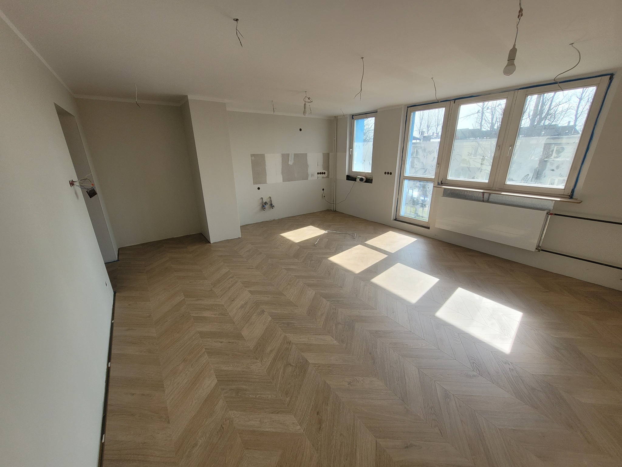 Wnętrze w trakcie prac wykończeniowych: ułożona podłoga w jodełkę, ściany przygotowane do malowania, widoczne instalacje elektryczne i hydrauliczne, okna z niebieską taśmą malarską.