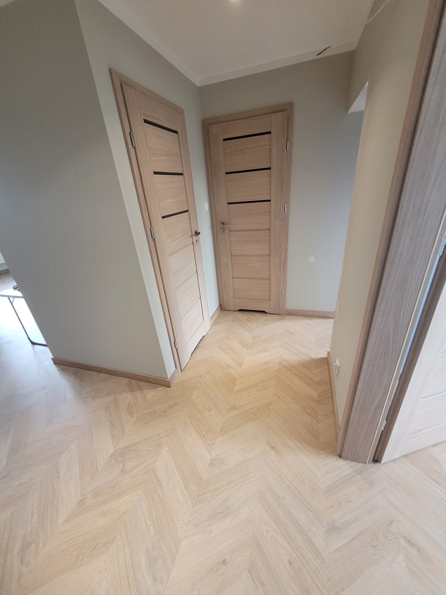 Jasny przedpokój z parkietem ułożonym we wzór jodełki, widoczne dwie drewniane, nowoczesne drzwi z czarnymi akcentami oraz fragment trzecich drzwi po prawej stronie.