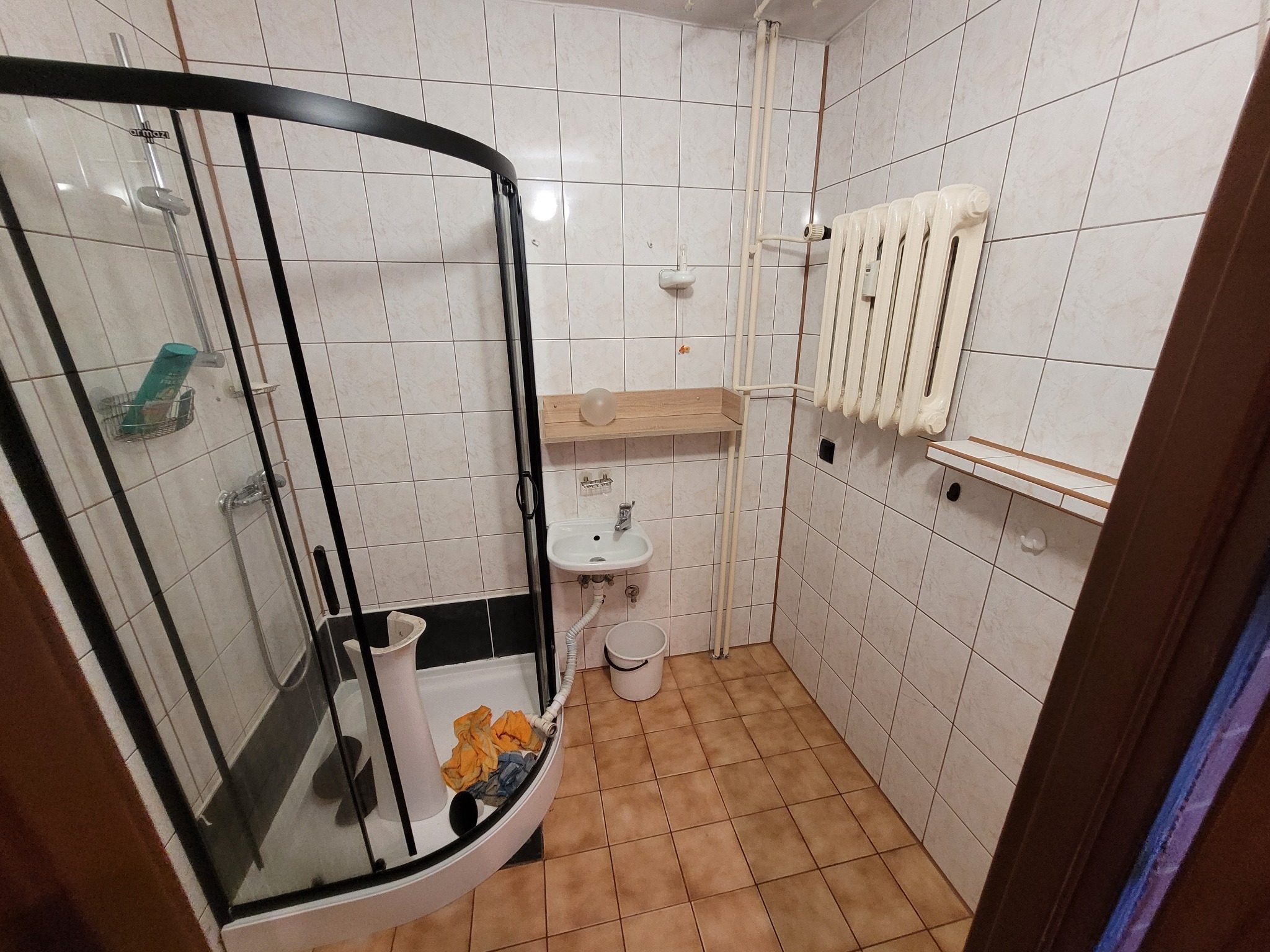 Łazienka z kabiną prysznicową z czarną ramą, umywalką na ścianie, grzejnikiem żeliwnym i beżowymi płytkami przed renowacją, widoczne narzędzia i materiały przygotowane do pracy.