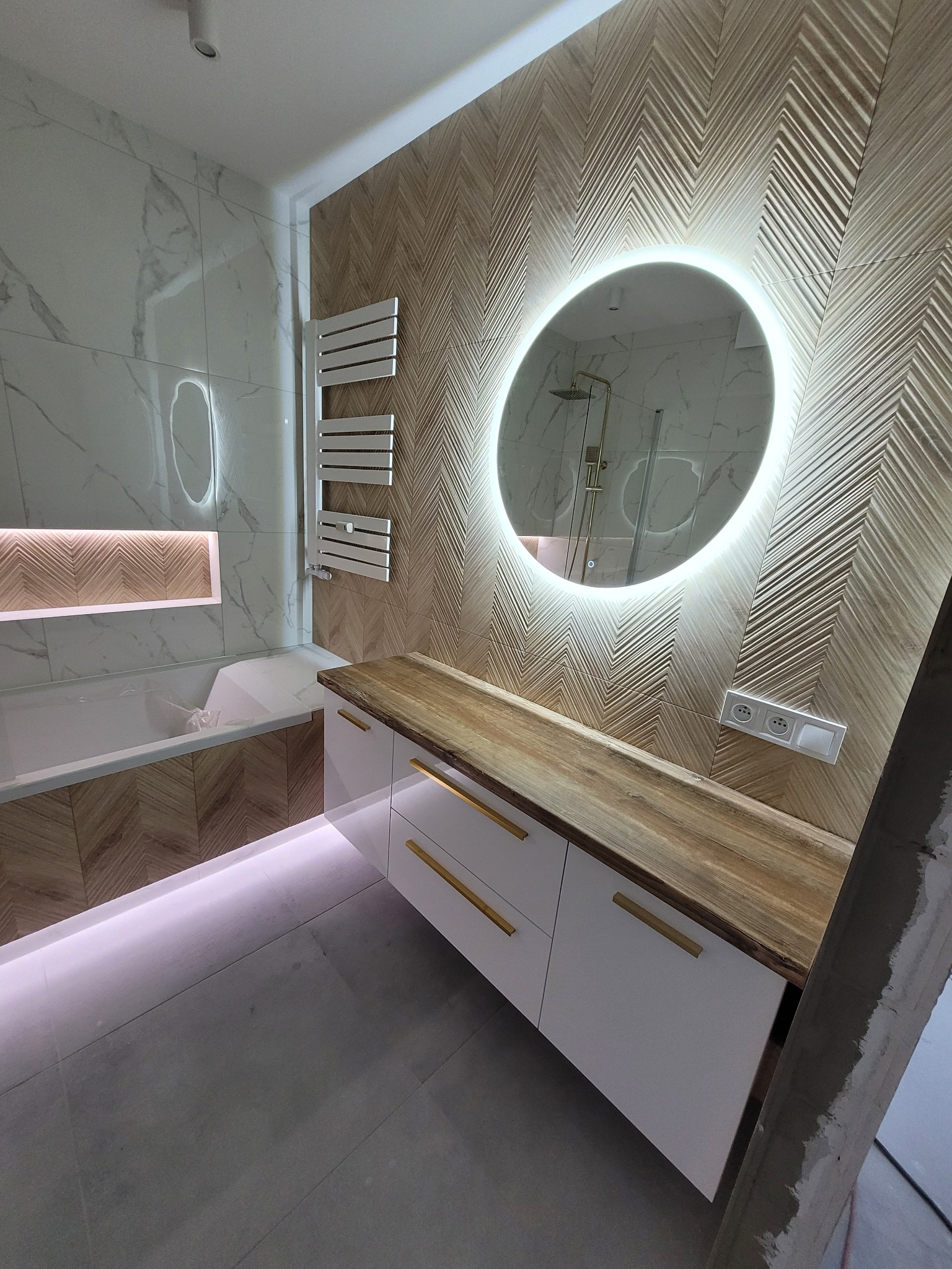 Wykończona łazienka z wzorzystą okładziną ścienną w jodełkę, okrągłym lustrem z podświetleniem LED, białą szafką z drewnianym blatem i złotymi uchwytami, oraz grzejnikiem drabinkowym.