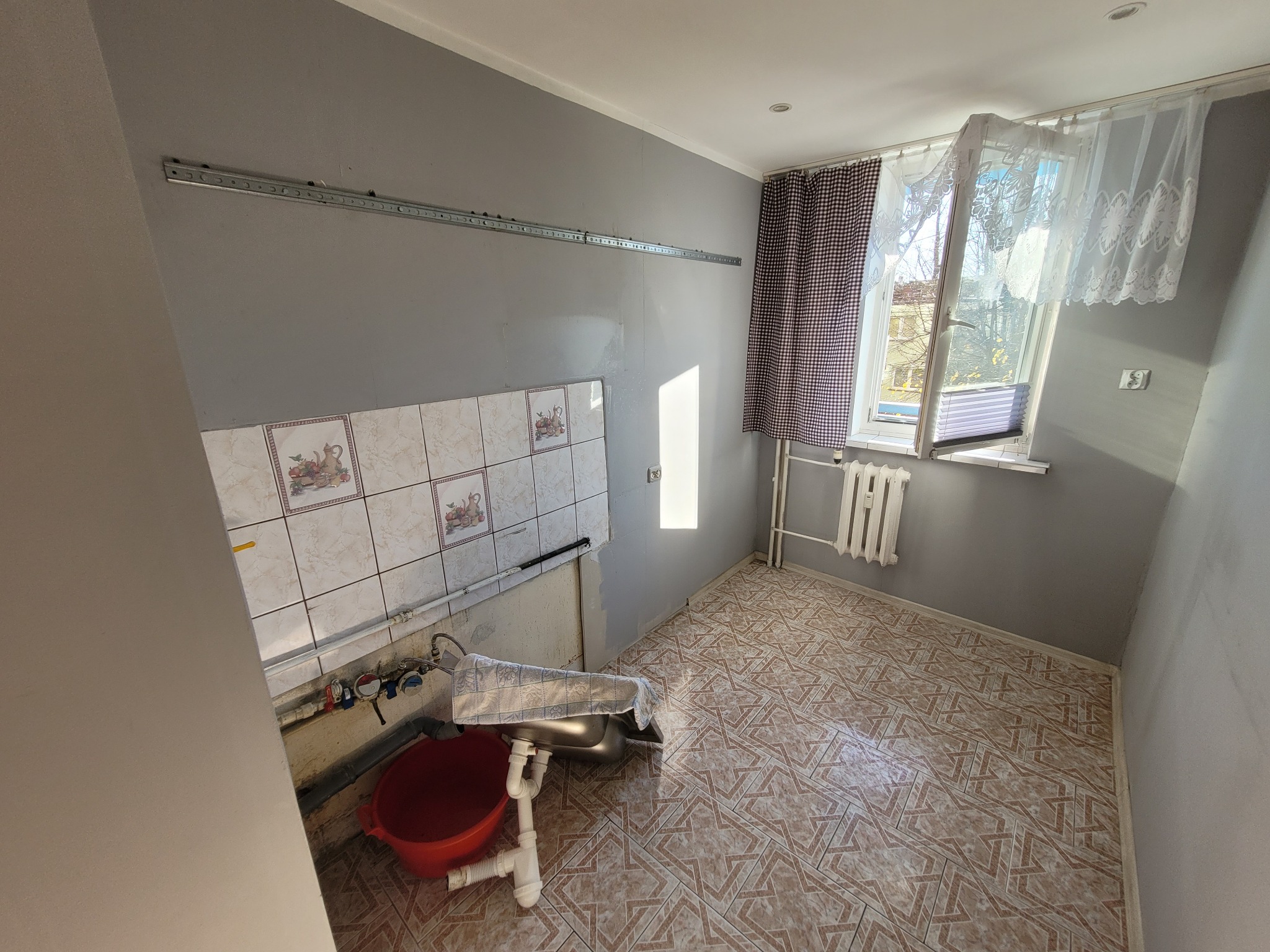 Kuchnia w trakcie remontu: widoczne płytki ścienne z motywem owoców, prowizorycznie zamontowany zlew przykryty szmatką, czerwone wiadro, szare ściany z zamontowanym profilem metalowym, brązowe...