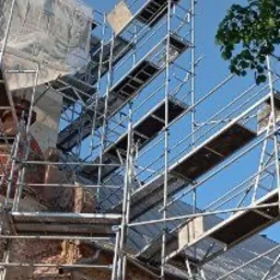 Wysokie rusztowanie z widocznymi platformami roboczymi ustawione przy elewacji budynku w trakcie prac remontowych, fragment zieleni drzew na tle błękitnego nieba.