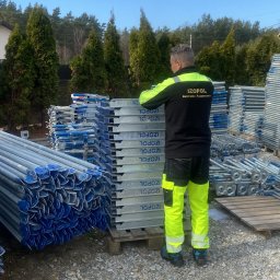 BELLAVIVRE - Pracownik w odzieży roboczej firmy Izopol stoi obok stosu elementów rusztowań na żwirowym podłożu, w tle iglaki i błękitne niebo. Widoczne elementy rusztowań.