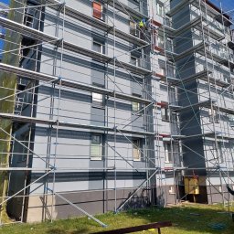 BELLAVIVRE - Szare, wielopiętrowe ocieplone rusztowanie elewacji budynku, z pracownikami na rusztowaniu i jednym na dole, w żółtej kamizelce, na tle błękitnego nieba.