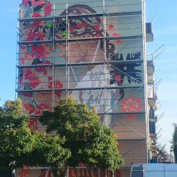 IZOPOL RUSZTOWANIA SPÓŁKA Z OGRANICZONĄ ODPOWIEDZIALNOŚCIĄ - Wielkoformatowy mural z motywem kobiety i kwiatów na ścianie bloku, widoczny z poziomu ulicy, z rusztowaniem i pracownikami w trakcie realizacji.
