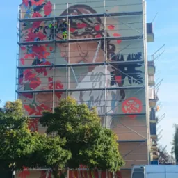 Wielkoformatowy mural z motywem kobiety i kwiatów na ścianie bloku, widoczny z poziomu ulicy, z rusztowaniem i pracownikami w trakcie realizacji.