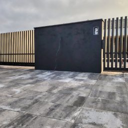 BELLAVIVRE - Nowoczesna, grafitowa brama posesji z pionowymi szczeblami, wykończona kostką brukową. Widoczne elementy instalacji elektrycznej na ścianie bramy.