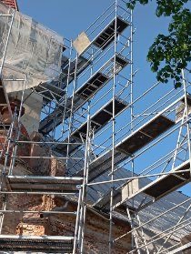 Wysokie rusztowanie z widocznymi platformami roboczymi ustawione przy elewacji budynku w trakcie prac remontowych, fragment zieleni drzew na tle błękitnego nieba.