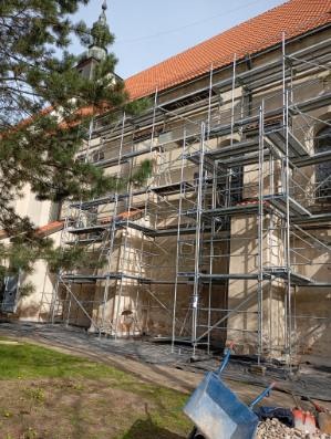Elewacja zabytkowego budynku z ceglanym dachem, częściowo zakryta rusztowaniem, widok z dołu, w tle drzewo iglaste i niebieskie taczki z gruzem.