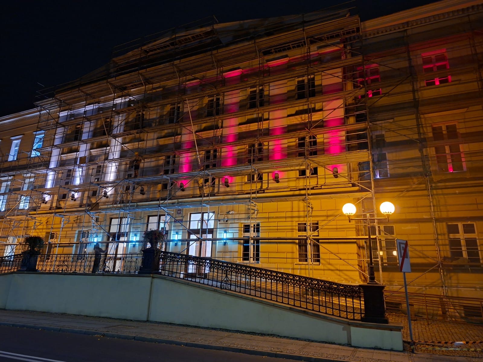 Elewacja budynku w rusztowaniach, oświetlona nocą kolorowymi światłami. Prace remontowe w toku, widoczne elementy konstrukcyjne i oświetlenie robocze.